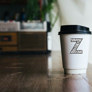 May include: A white takeaway coffee cup with a black lid. The cup has a black and white patterned letter "Z" on the front.