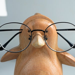 May include: A wooden penguin figurine wearing black round glasses. The glasses have a black frame and clear lenses.