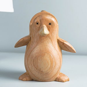 May include: A wooden penguin figurine with a light brown color and a gold beak. The penguin is standing on a white surface.