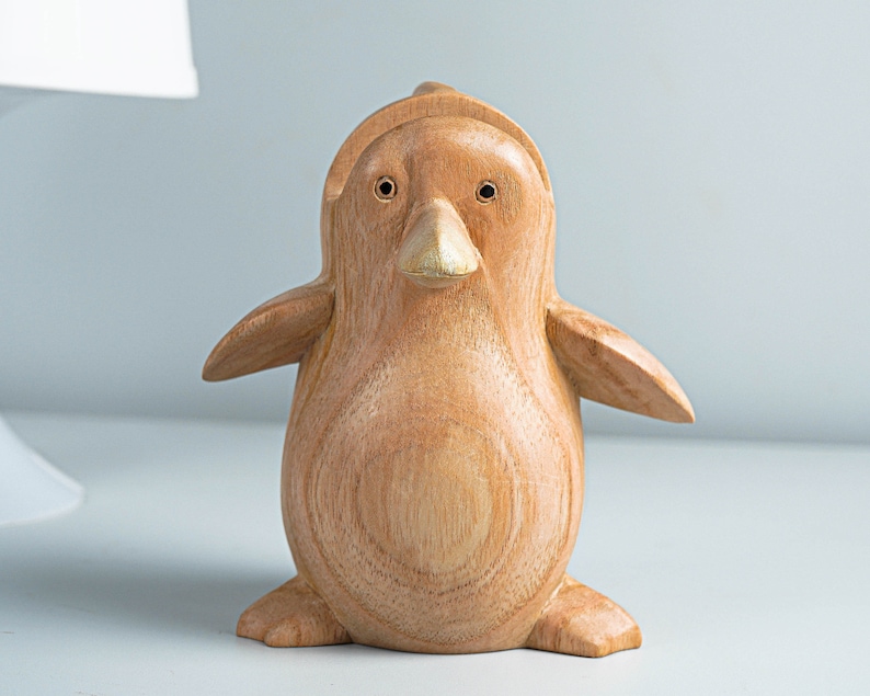 May include: A wooden penguin figurine with a light brown color and a gold beak. The penguin is standing on a white surface.