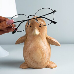 May include: A wooden penguin figurine with black rimmed glasses perched on its beak. The penguin is light brown with a smooth, slightly textured surface.