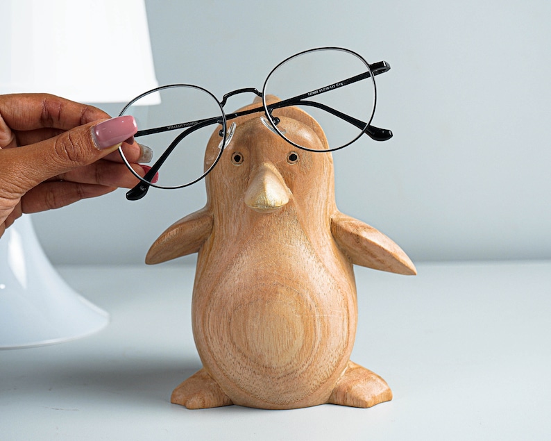 May include: A wooden penguin figurine with black rimmed glasses perched on its beak. The penguin is light brown with a smooth, slightly textured surface.