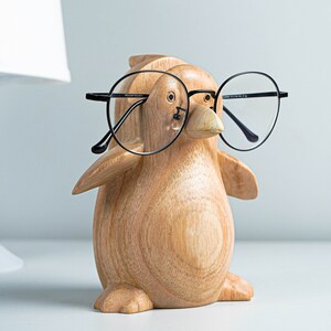 May include: A wooden penguin figurine wearing black framed glasses. The penguin is light brown with a detailed wood grain texture.