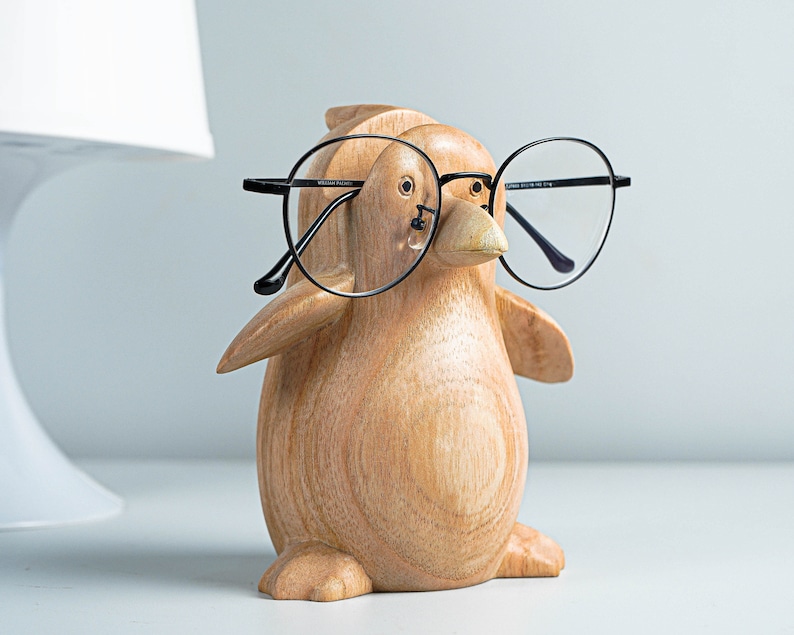 May include: A wooden penguin figurine wearing black framed glasses. The penguin is light brown with a detailed wood grain texture.