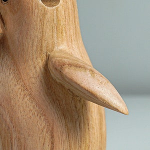 May include: A wooden penguin figurine with a smooth, light brown finish. The penguin has a rounded body and a long, curved beak. The figurine's wing is outstretched, showing the detailed wood grain.