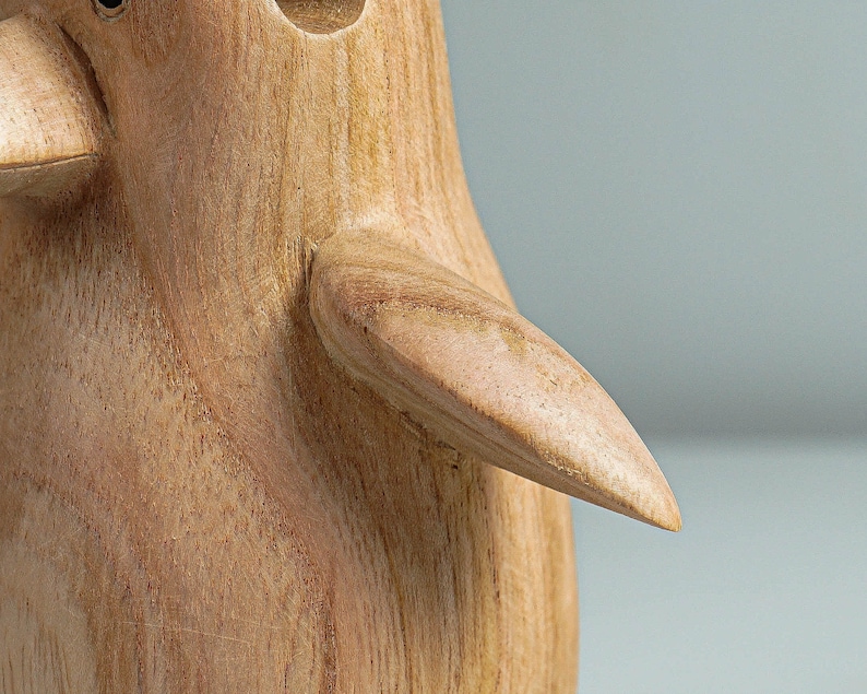 May include: A wooden penguin figurine with a smooth, light brown finish. The penguin has a rounded body and a long, curved beak. The figurine's wing is outstretched, showing the detailed wood grain.