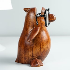 May include: A wooden penguin figurine with black glasses, a brown finish, and a carved wing. The penguin is standing on a white surface.