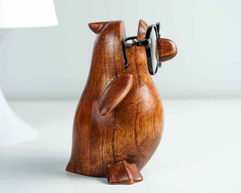 May include: A wooden penguin figurine with black glasses, a brown finish, and a carved wing. The penguin is standing on a white surface.