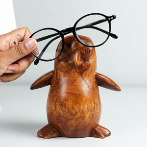 May include: A wooden penguin figurine with a dark brown finish, holding a pair of black eyeglasses.
