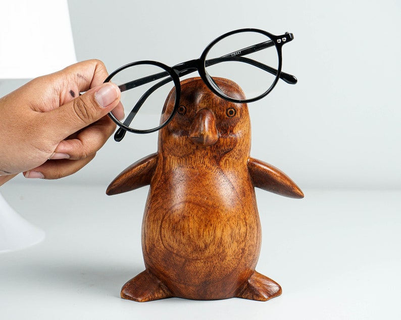 May include: A wooden penguin figurine with a dark brown finish, holding a pair of black eyeglasses.