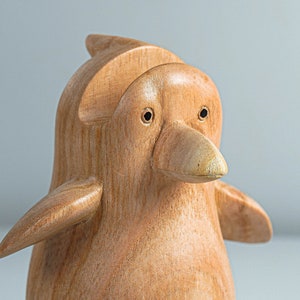 May include: A wooden penguin figurine with a light brown color. The penguin has a small beak and two black eyes.