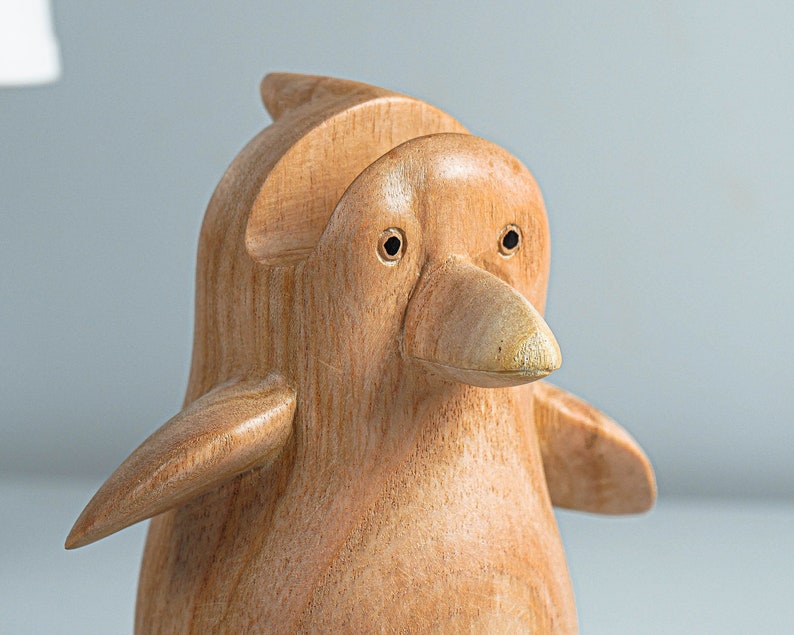 May include: A wooden penguin figurine with a light brown color. The penguin has a small beak and two black eyes.