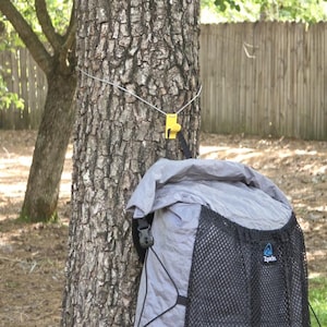 Può includere: Uno zaino grigio con chiusura a rullo, tasca frontale in rete nera e logo Zpacks, appeso al tronco di un albero tramite un cordino bianco fissato a un gancio in plastica giallo brillante che sostiene lo zaino contro la corteccia ruvida.