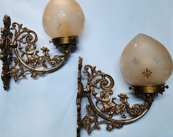 May include: Two antique wall sconces with ornate brass frames and frosted glass shades. Each sconce features a cherub design and star pattern on the glass. The sconces are designed to be mounted on a wall.