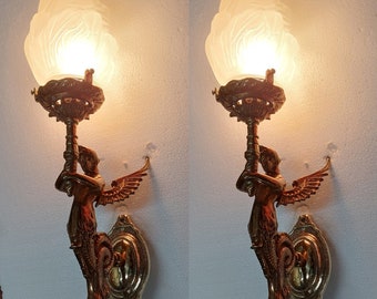 May include: Two gold-toned wall sconces with frosted glass shades and a winged cherub design. Each sconce features a decorative base and a single light bulb.