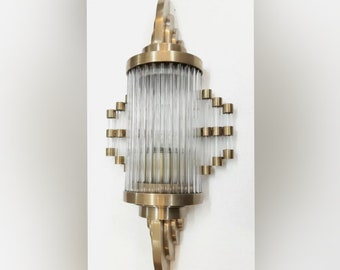 May include: A gold-toned wall sconce with a clear glass shade featuring vertical lines. The sconce has a decorative design with a curved top and bottom.