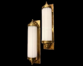 May include: Two gold wall sconces with white frosted glass shades. The sconces have a cylindrical shape and are decorated with vertical lines.