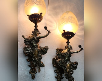 May include: A pair of antique brass wall sconces with frosted glass shades. Each sconce features a sculpted figure of a woman holding a musical instrument.