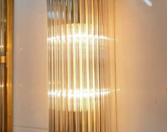 May include: A cylindrical wall sconce with a brass finish and a clear glass tube design. The light fixture has a warm glow, and the glass tubes are arranged vertically. The sconce is mounted on a white tiled wall.
