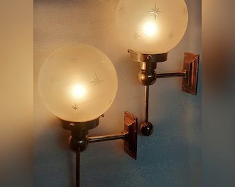 Può includere: Due applique vintage con globi in vetro smerigliato decorati con motivi a stella. Le applique hanno elementi in metallo color ottone e sono montate a parete. I globi sono illuminati, emettendo una calda luce.