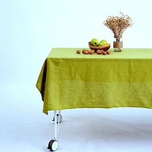 May include: A green linen tablecloth draped over a white table with wheels. The table is set with a wooden bowl of green apples and walnuts.