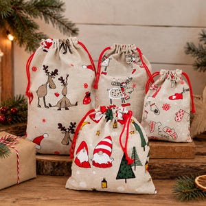 Könnte beinhalten: Vier beigefarbene Stoffbeutel mit Kordelzug und weihnachtlichen Motiven. Die Beutel zeigen Illustrationen von Rentieren, Gnomen, Tannenbäumen und Fäustlingen in Rot-, Grün- und Brauntönen. Jeder Beutel ist oben mit leuchtend roten Kordeln verschlossen.