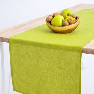 May include: A green linen table runner with a wooden bowl filled with green apples and walnuts on a wooden table.