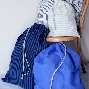 Peut inclure: Trois sacs à linge en lin rayés. Un sac est bleu et blanc, un autre est beige et blanc, et le dernier est bleu. Les sacs sont posés sur une chaise en bois.