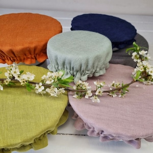 Peut inclure: Plusieurs couvercles de bols ronds en tissu de différentes couleurs : orange, vert, bleu, gris et rose. Chaque couvercle a un bord à volants. Une branche fleurie avec des fleurs blanches est posée sur les couvercles.