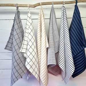 May include: Six kitchen towels hanging on a wooden dowel. The towels are made of linen and have different patterns, including stripes, checks, and a plain colour. The towels are all in neutral colours, such as grey, white, and beige.