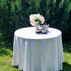 May include: A white tablecloth with a black checkered pattern covers a round table set for tea. A vase with white flowers and a teapot with two teacups are on the table.
