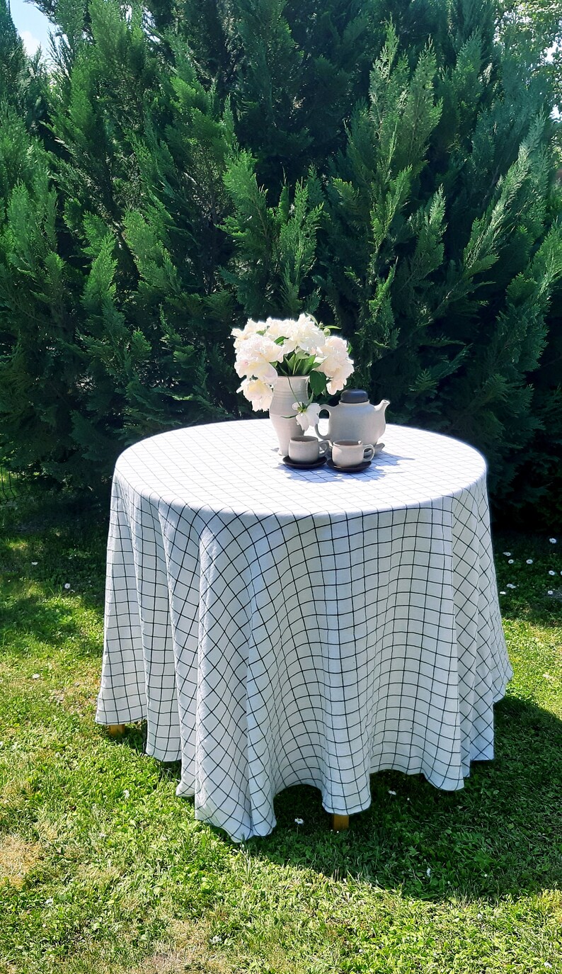 May include: A white tablecloth with a black checkered pattern covers a round table set for tea. A vase with white flowers and a teapot with two teacups are on the table.