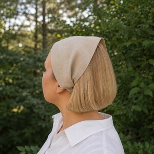 May include: A beige linen head covering, tied at the back, worn over short blonde hair. The head covering is paired with a white collared shirt. The background features green foliage, suggesting an outdoor setting. The head covering is a neutral color.