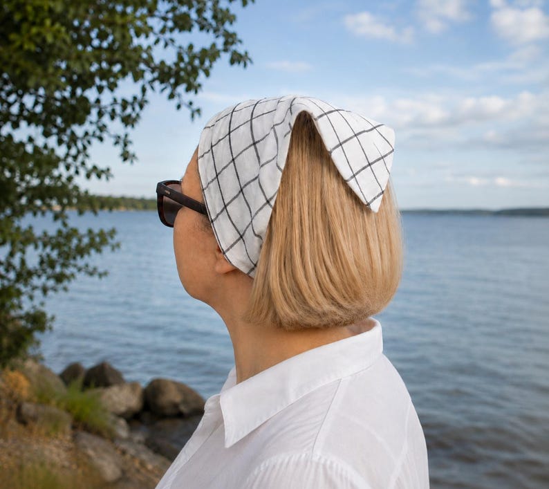 May include: A white bandana with a black grid pattern is worn on the head. The person is wearing sunglasses and a white collared shirt. The background shows a body of water and a blue sky.
