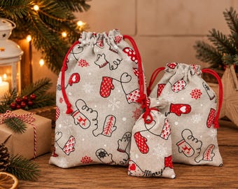 May include: Three beige drawstring gift bags of varying sizes featuring a repeating pattern of red and white mittens, snowflakes, and circular motifs. Each bag has bright red cord drawstrings and the smallest bag has the word Merry printed.