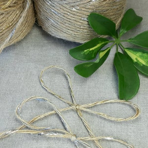 May include: Two spools of natural jute twine with silver metallic accents. The twine is tied in a bow on a light gray linen fabric background.