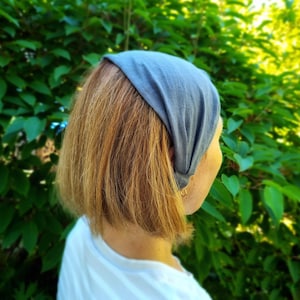 May include: A gray headband worn on the head of a person with short, light brown hair. The headband is made of a soft fabric and is worn over the forehead. The background is a lush green bush.