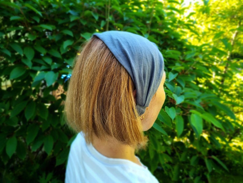 May include: A gray headband worn on the head of a person with short, light brown hair. The headband is made of a soft fabric and is worn over the forehead. The background is a lush green bush.