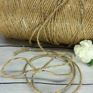 May include: A roll of natural jute twine with silver metallic thread running through it. The twine is unraveled and laid out on a white wooden surface.