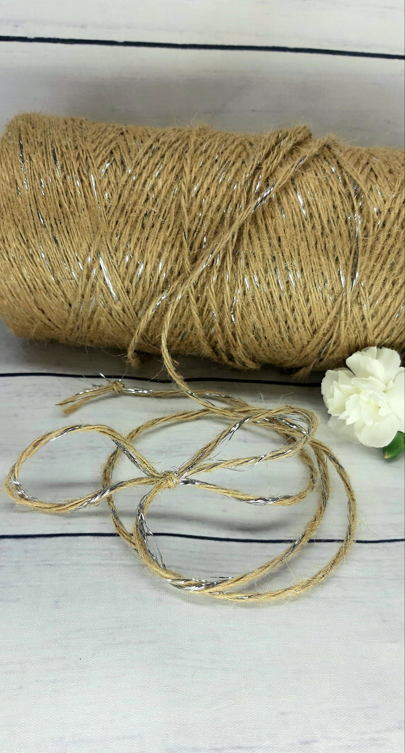 May include: A roll of natural jute twine with silver metallic thread running through it. The twine is unraveled and laid out on a white wooden surface.