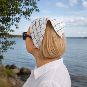 May include: A white bandana with a black grid pattern is worn on the head. The person is wearing sunglasses and a white collared shirt. The background shows a body of water and a blue sky.