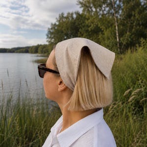 May include: A woman wearing a light beige headscarf and sunglasses, looking towards a lake. The headscarf is folded and tied at the back. She is wearing a white collared shirt. The background features a lake, trees, and tall grass.