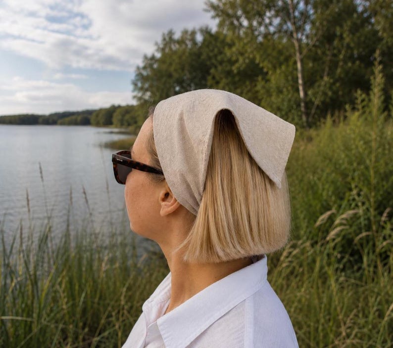 May include: A woman wearing a light beige headscarf and sunglasses, looking towards a lake. The headscarf is folded and tied at the back. She is wearing a white collared shirt. The background features a lake, trees, and tall grass.