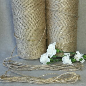 May include: Two spools of natural jute twine with gold glitter accents. The twine is wound around the spools and is ready to be used for crafting or decorating. There are also several strands of the twine lying on a light gray surface.