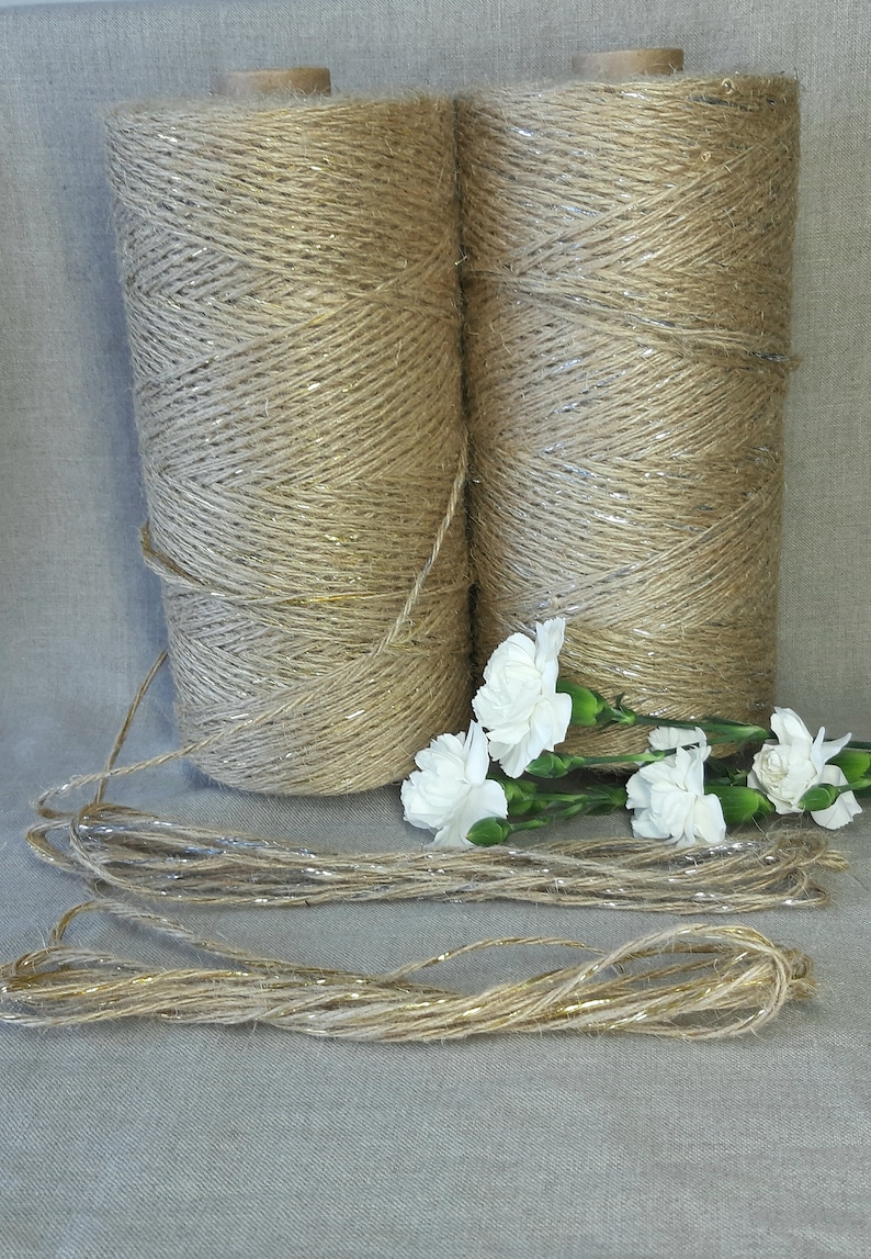 May include: Two spools of natural jute twine with gold glitter accents. The twine is wound around the spools and is ready to be used for crafting or decorating. There are also several strands of the twine lying on a light gray surface.