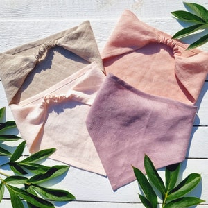 May include: Four linen headbands in shades of beige, pink, and purple are arranged on a white wooden surface. The headbands have a twisted knot design and an elastic band at the back. Green leaves are scattered around the headbands.