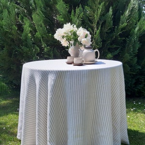May include: A white linen tablecloth with gray stripes, set on a round table outdoors. A bouquet of white flowers and a teapot with two cups and saucers are on the table.