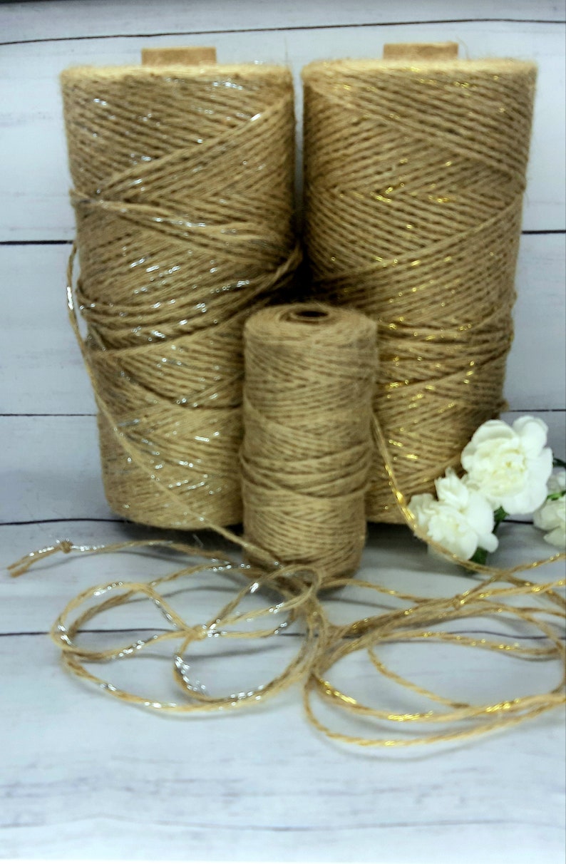 May include: Three spools of twine with gold and silver accents. The twine is brown and the spools are made of cardboard. The twine is lying on a white wooden surface.