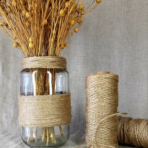 May include: A glass vase wrapped in natural twine with dried flax flowers inside. Two spools of twine are also shown on a linen background.