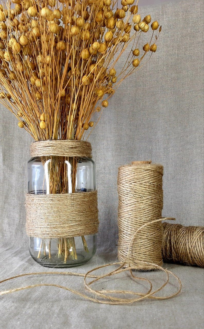 May include: A glass vase wrapped in natural twine with dried flax flowers inside. Two spools of twine are also shown on a linen background.